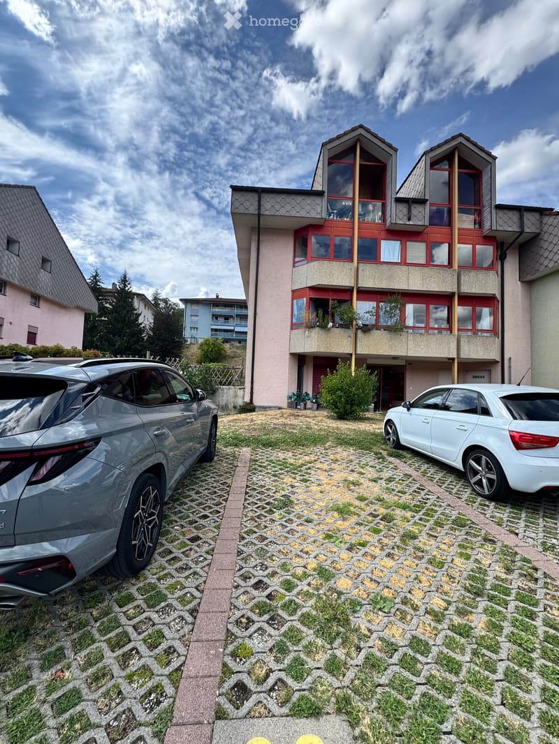 Modern two-story house with red-framed windows, balconies, and a paved driveway with two parked cars.