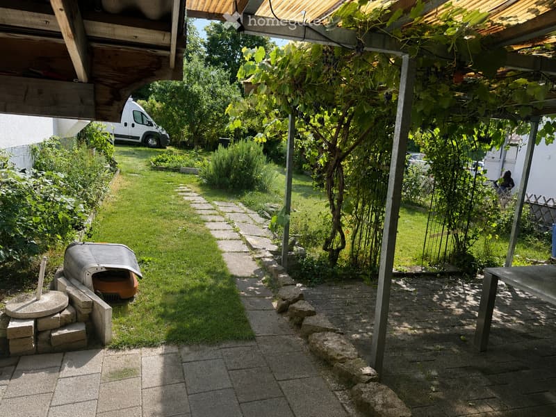 Garden area with a stone pathway, grassy lawn, covered patio with vines, and a barbecue grill.