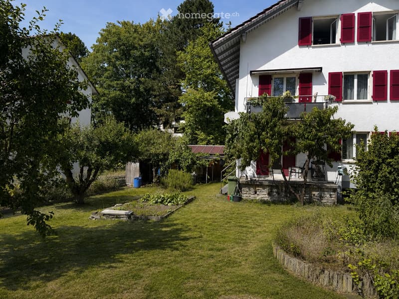 Two-story white house with red shutters, a balcony, and a well-maintained garden with grass, trees, and shrubs.