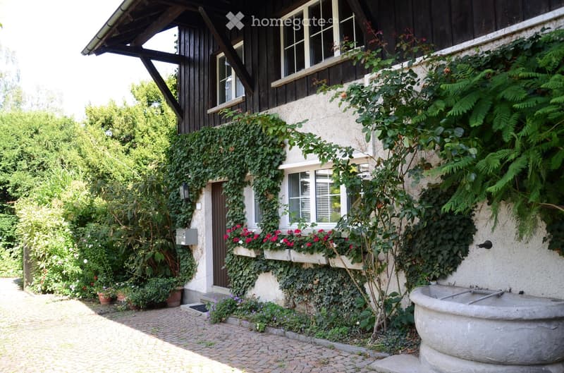 A two-story house with a stone facade and wooden upper section, covered in climbing plants and flowers, with a brick pathway leading to the entrance.