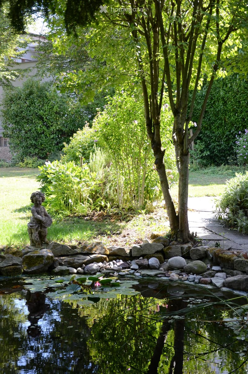 A serene garden with a pond surrounded by rocks, lily pads, lush greenery, and a small statue on a stone platform.