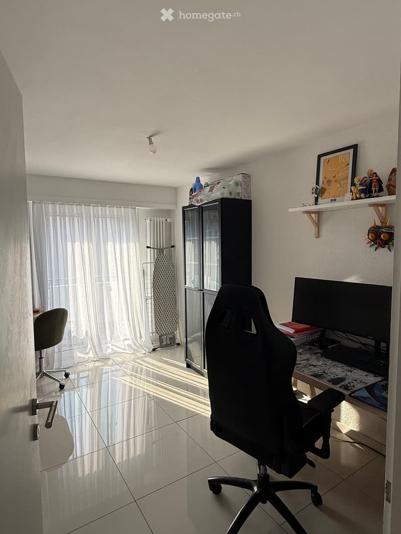 A furnished room with a desk, chair, screen, keyboard, shelf, and cabinet. The room has a tiled floor and a window with white curtains.