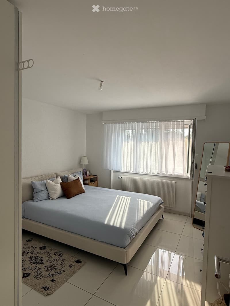 Bright bedroom with a large bed, nightstand, lamp, mirror, and radiator. The room features tiled flooring and a patterned rug.