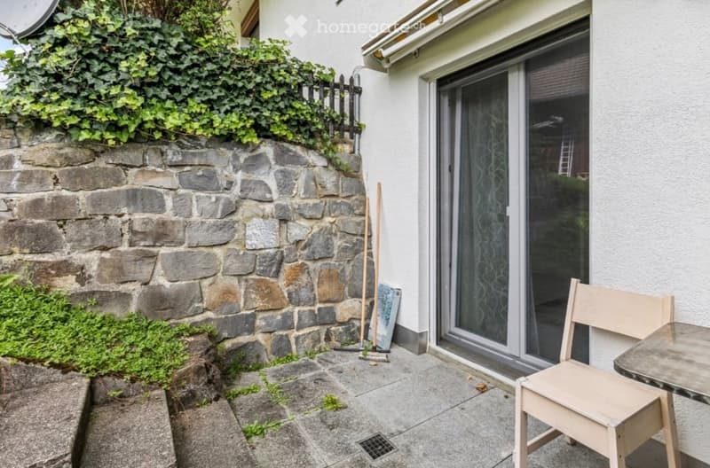 Small outdoor patio area with a stone wall, wooden chair, small table, broom, and a sliding glass door leading to the house.