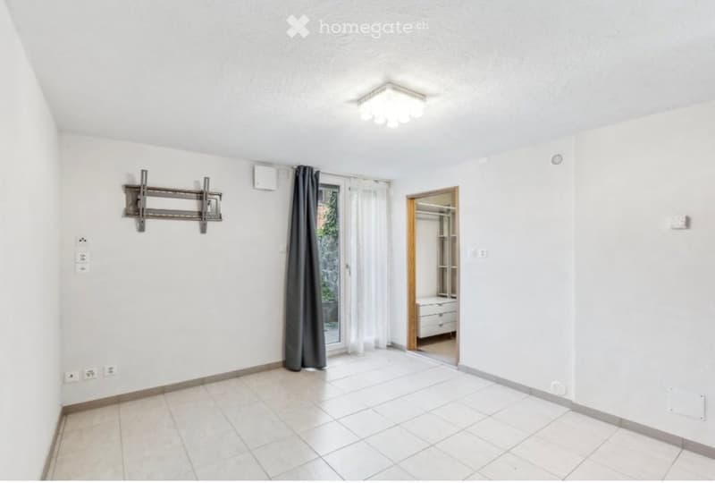 Empty room with white walls, tiled floor, and a sliding glass door leading to a balcony. A TV mount is attached to the wall, and a small closet with drawers is visible in the adjacent space.