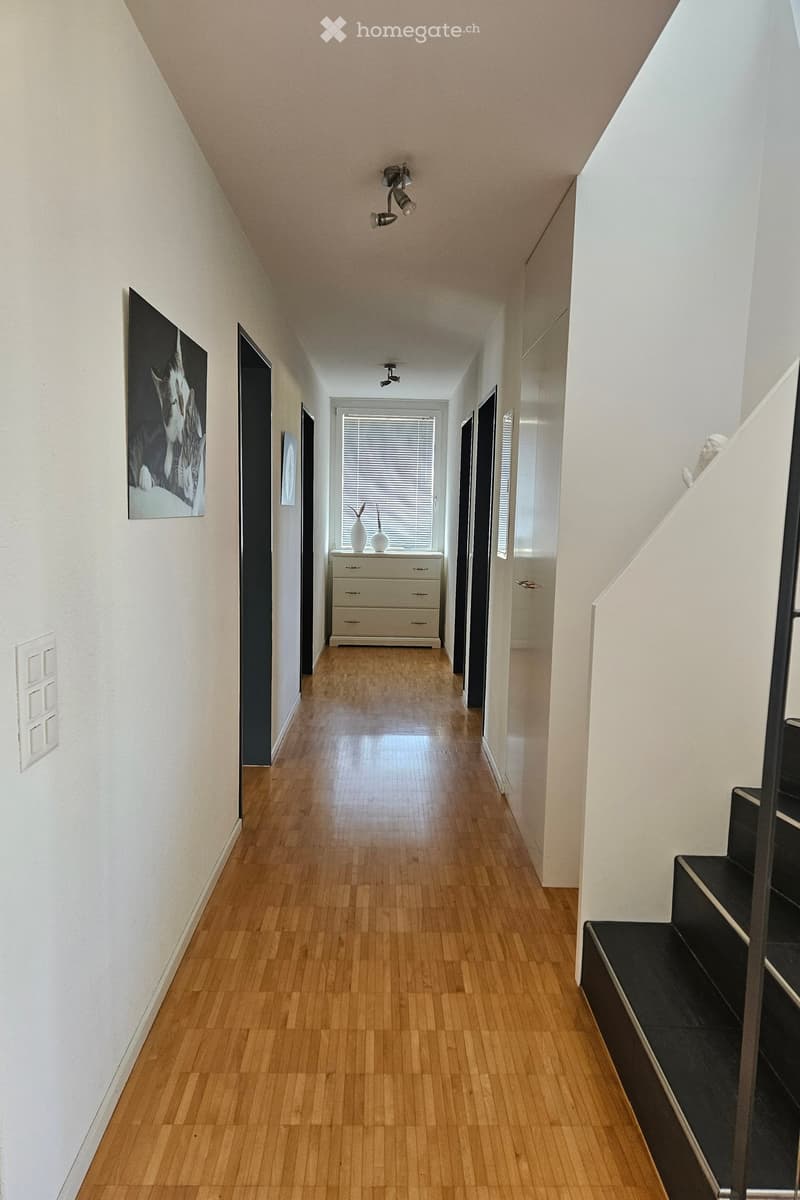 A long, narrow hallway with wooden flooring, white walls, and modern lighting fixtures. A staircase leads upwards on the right, and a small room with a dresser and vases is visible at the end.