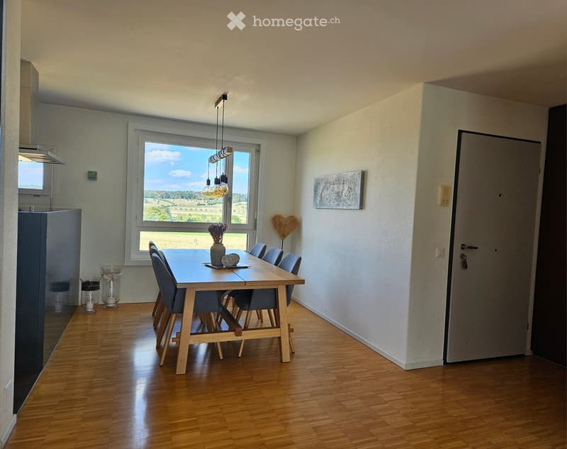 Bright dining room with wooden floor, large window overlooking a landscape, dining table with chairs, decorative items including a vase and candles.