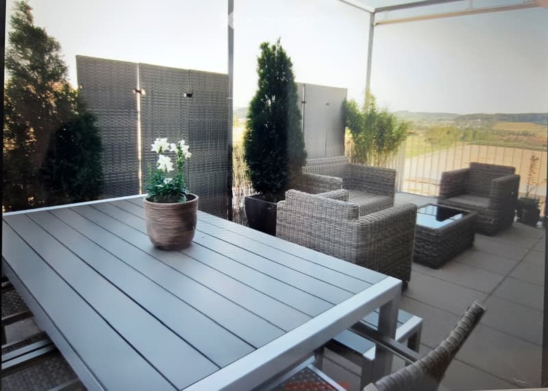 Balcony with a grey wooden table, wicker chairs, sofa, and potted plants, offering a scenic view.
