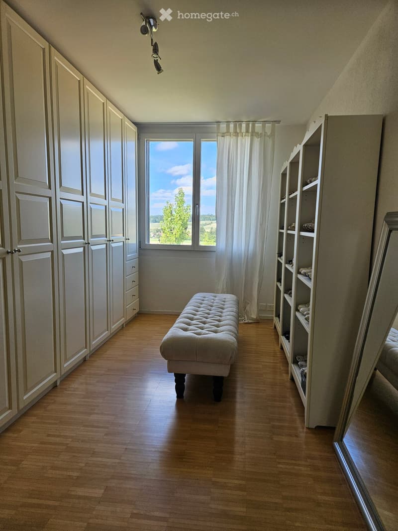 Bright room with wooden flooring, large window with white curtains, built-in wardrobe, and a small bench or stool in the center.