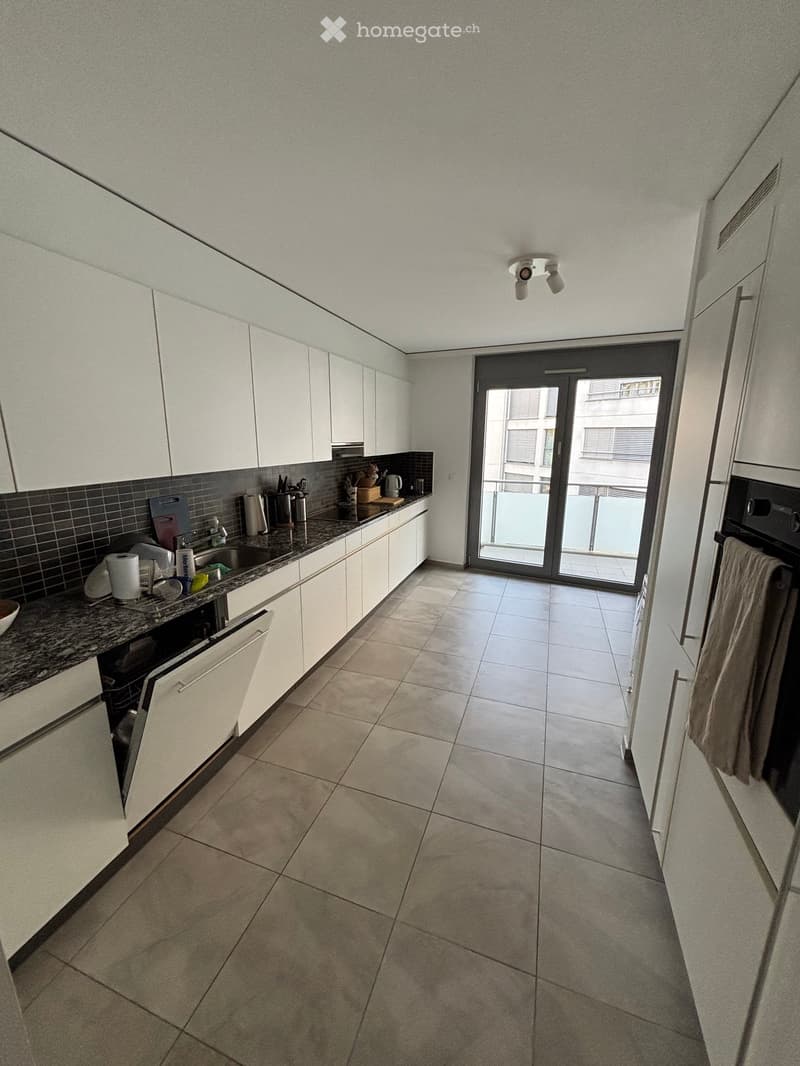Modern kitchen with white cabinets, granite countertop, and tiled floor. Features a sink, dishwasher, and oven, with a sliding glass door leading to a balcony.