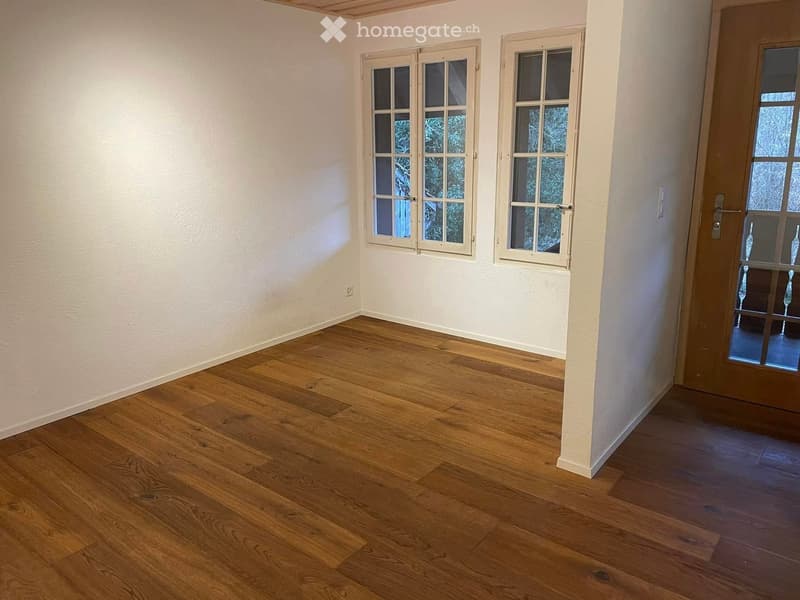 Empty room with wooden flooring, white walls, and large windows with white frames. A door with glass panels is visible on the right side.