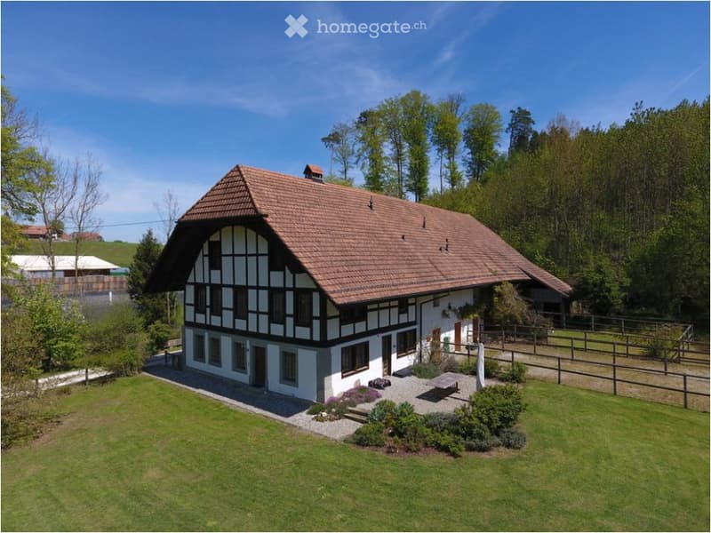 A two-story house with a traditional half-timbered design, red-tiled roof, and white walls. The house is surrounded by a well-maintained lawn, small garden beds, and a wooden fence.