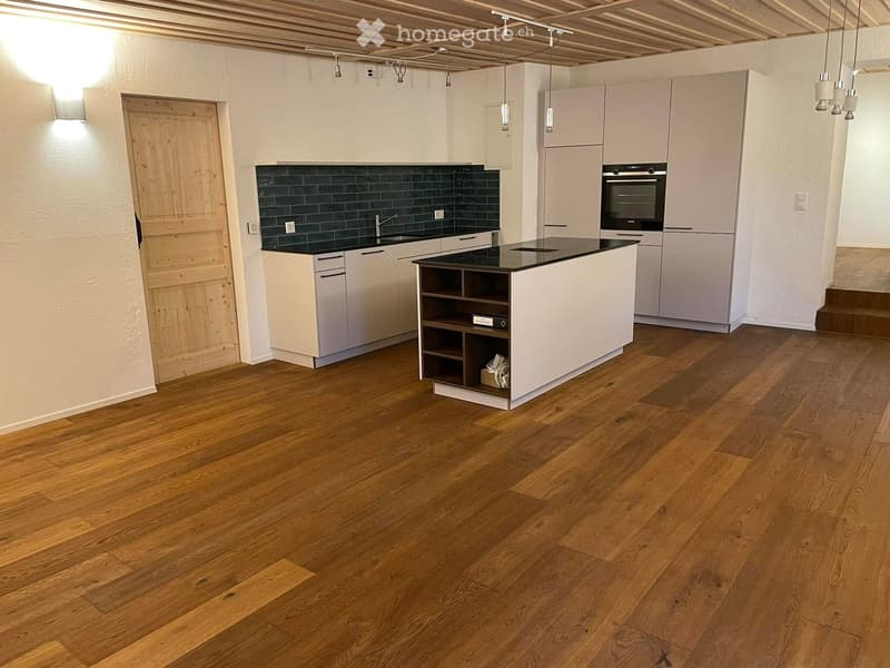Modern kitchen with light-colored cabinets, a central island, and a dark green backsplash. The floor is made of light wooden planks.