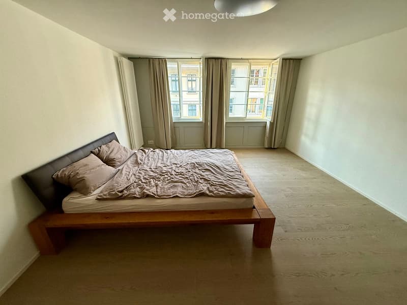 A modern bedroom with a wooden bed, beige bedding, and large windows with curtains. The room features light-colored walls and a wooden floor.