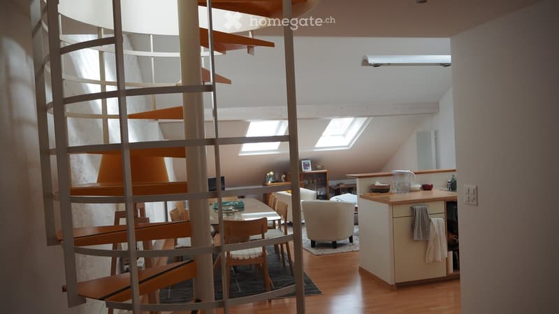 Modern, open-plan living and dining area with a spiral staircase, wooden flooring, and large windows. Features a sofa, dining table with chairs, and a small kitchen counter.