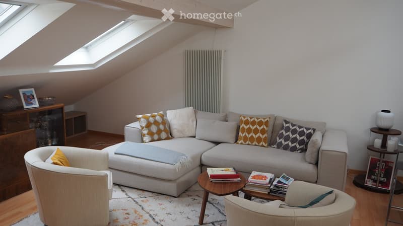 Modern living room with a grey sectional sofa, colorful throw pillows, a coffee table with books, two armchairs, a wooden side table, and a radiator. The room features wooden flooring, a patterned rug, and skylight windows.