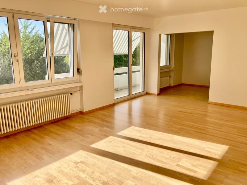 Empty room with light wooden flooring, large windows with blinds, and radiators along the walls. The space is unfurnished and appears to be a living or dining area.