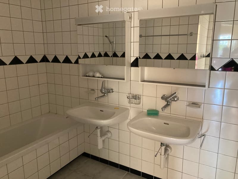 Bathroom with two sinks, a bathtub, mirrors, shelves, and a small window.