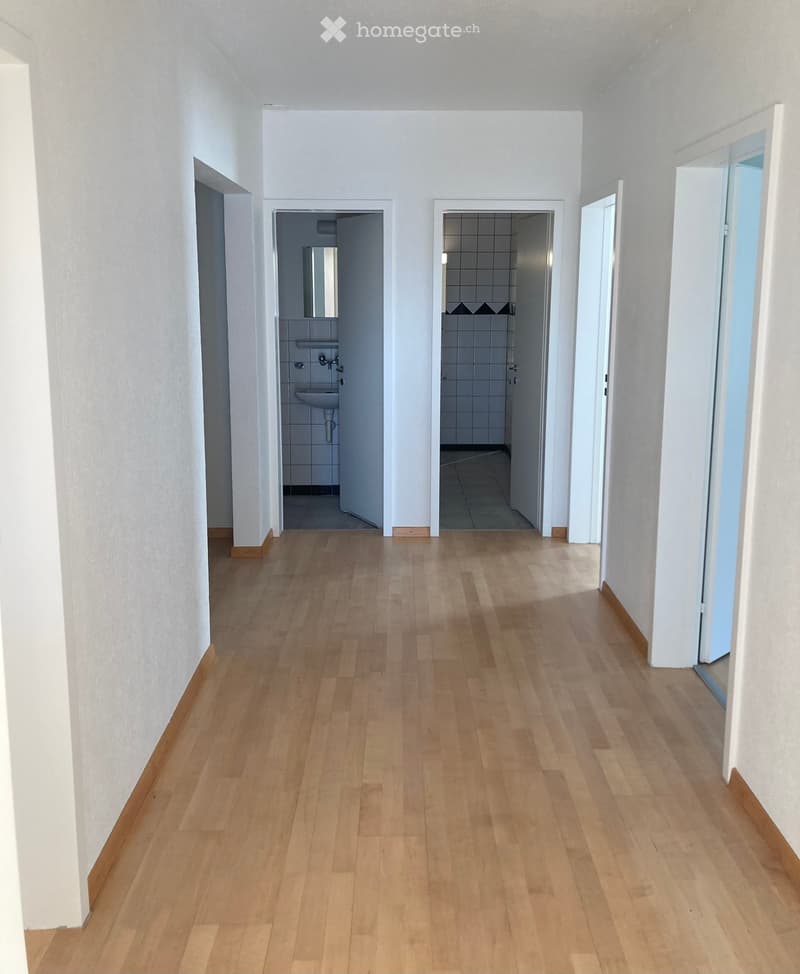 Empty hallway with light wooden flooring and white walls, leading to two adjacent rooms: a bathroom and a shower area.