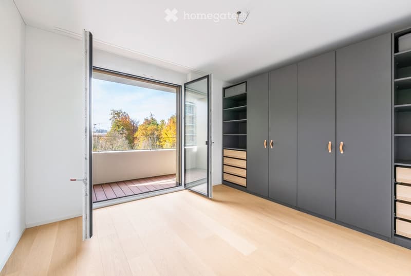 Empty room with light wooden flooring, white walls, large sliding glass doors leading to a balcony with a view of trees and buildings.