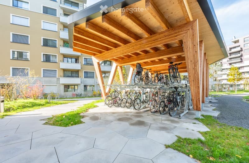 Outdoor bike parking area with wooden shelter, multiple bicycles, and modern building in the background.