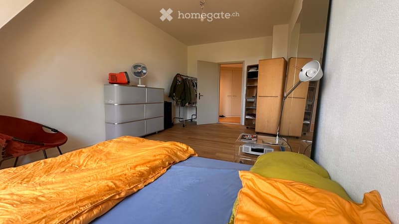 Cozy bedroom with a bed, a red chair, a wardrobe, a fan, a lamp, and a small table. The room has wooden flooring and white walls.