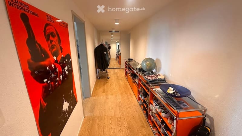 A long, narrow hallway with wooden flooring, white walls, and a large red poster on the left side. A shoe rack with multiple pairs of shoes is visible on the right, along with a small globe and some folded clothes. The hallway leads to an open doorway at