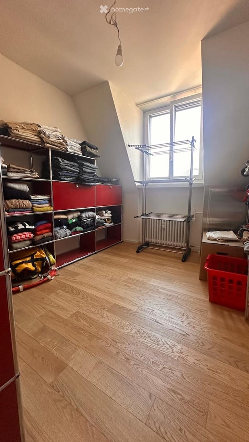A room with wooden flooring, a large window, a shelf filled with folded clothes, a radiator, a clothes hanger, and a red basket on the floor.