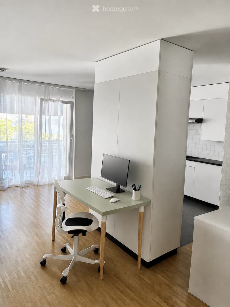 A modern, minimalist office space with a light green desk, ergonomic chair, computer monitor, keyboard, mouse, and a cup on the desk. The room features light wooden flooring and white cabinetry.