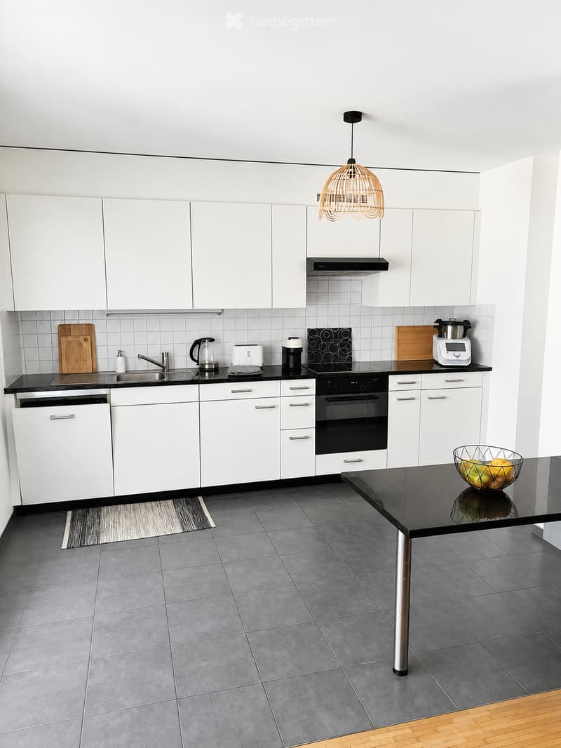 Modern kitchen with white cabinets, black countertops, stainless steel appliances, and a central island with a bowl of fruit.