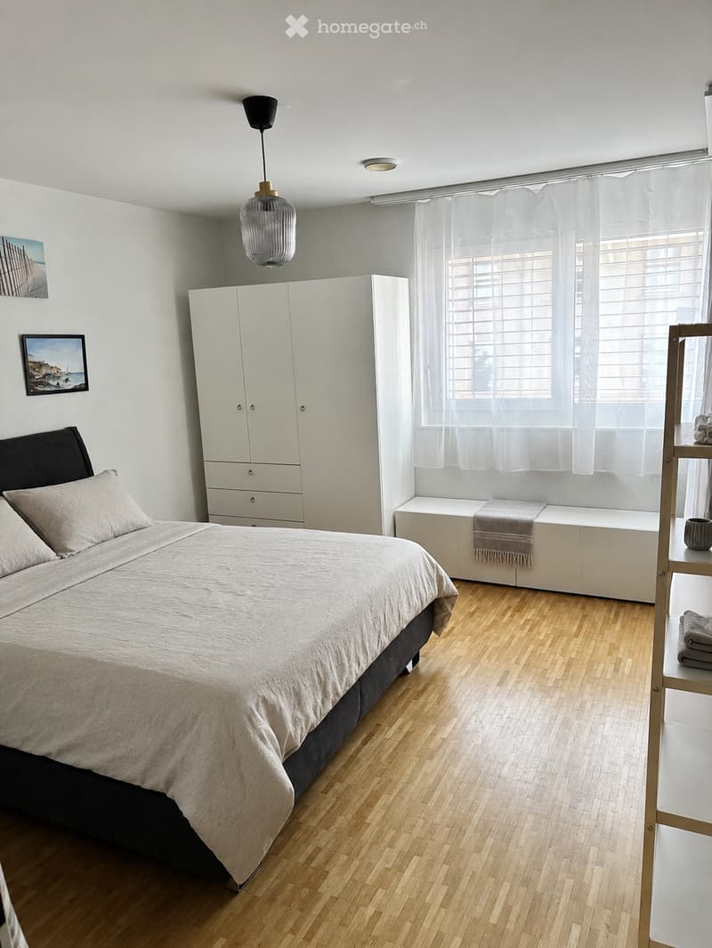 Modern bedroom with a large bed, white wardrobe, wooden floor, large window with white curtains, and a bench with a towel.