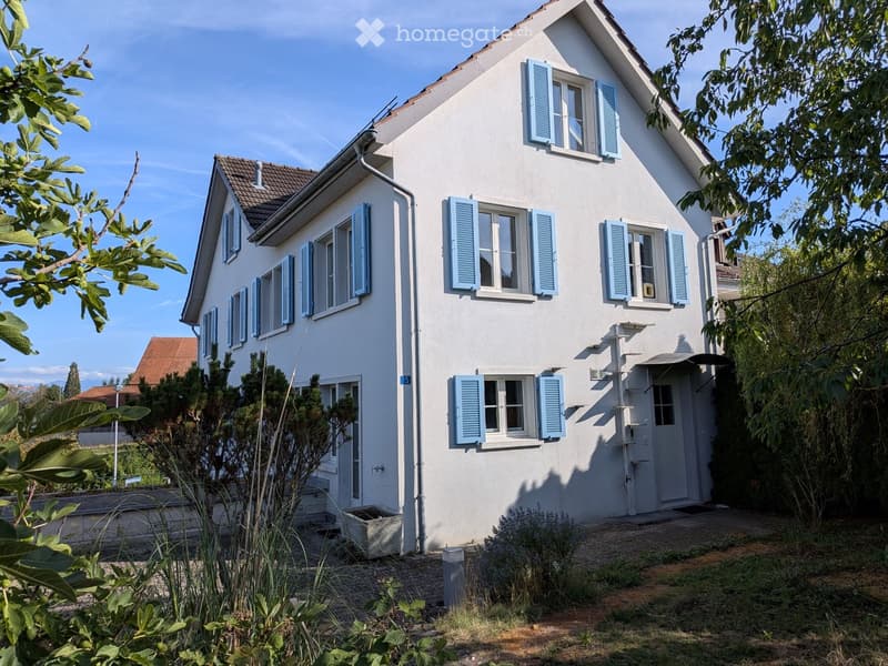 Two-story white house with blue shutters, surrounded by greenery and a small garden area.