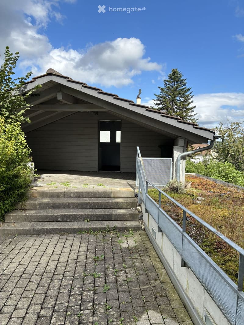 A small, modern building with a sloped roof and a covered entrance, surrounded by greenery and a paved area.
