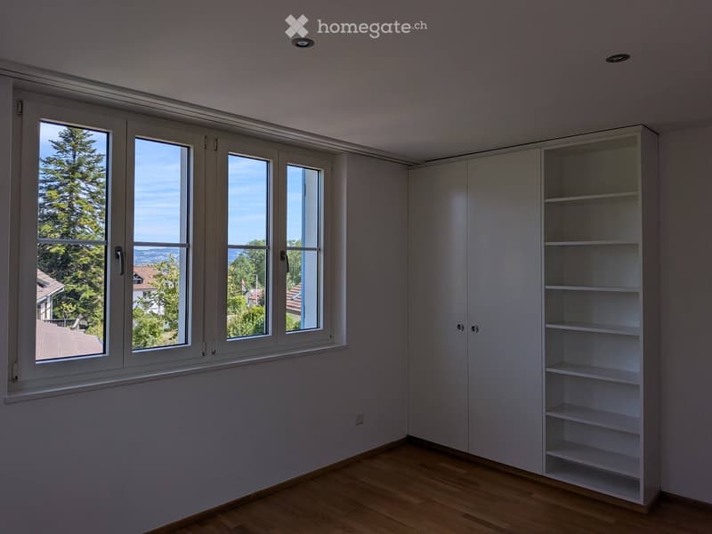 Empty room with wooden flooring, white walls, large windows with white frames, and built-in white shelving unit.