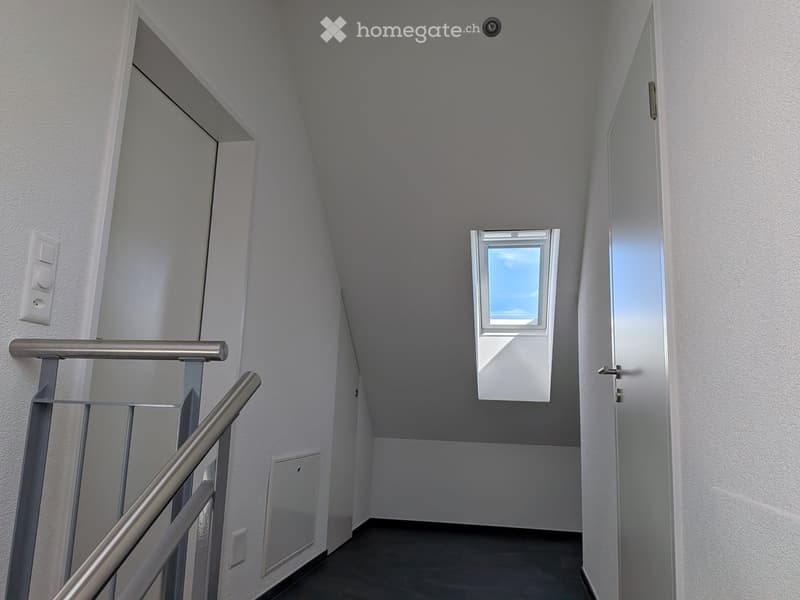 A narrow hallway with white walls, a metal railing on the left, and a small window at the end, allowing natural light to enter.