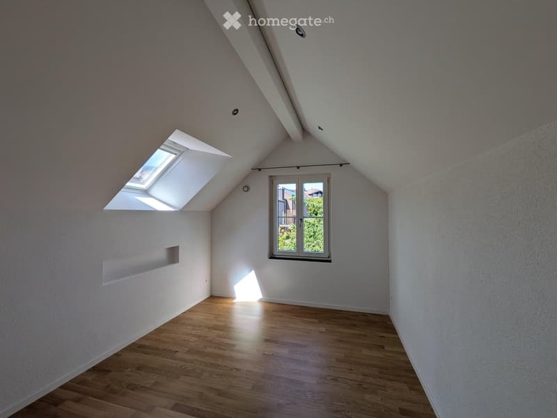 Empty attic room with wooden flooring, white walls, skylight window, and a regular window with a view of greenery.