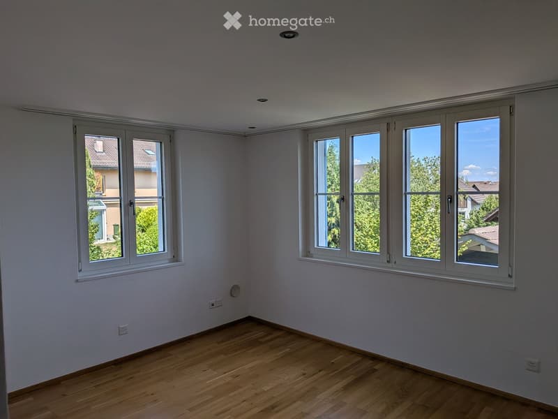 Empty room with light grey walls, wooden floor, and large windows offering a view of neighboring houses and greenery.