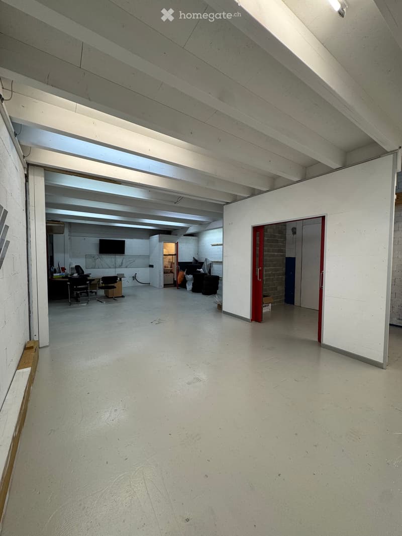 Open, industrial-style room with high ceilings, exposed beams, and white walls. Contains a few chairs, a table, a screen, and a refrigerator in the background.