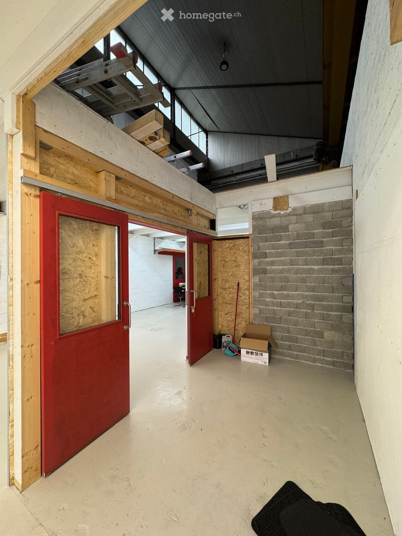 Empty hallway with exposed wooden beams, concrete floors, and red doors leading to other rooms. Visible construction materials and storage items are present.