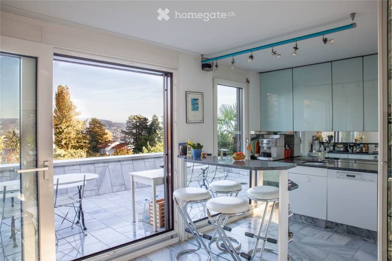 Modern kitchen with bar counter, stools, and a view of the landscape through large windows.