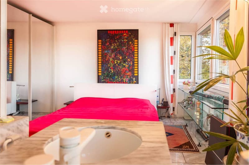 Bright bedroom with a red bedspread, a nightstand, a sink, a mirror, a glass table, a chair, a plant, and a colorful painting on the wall.