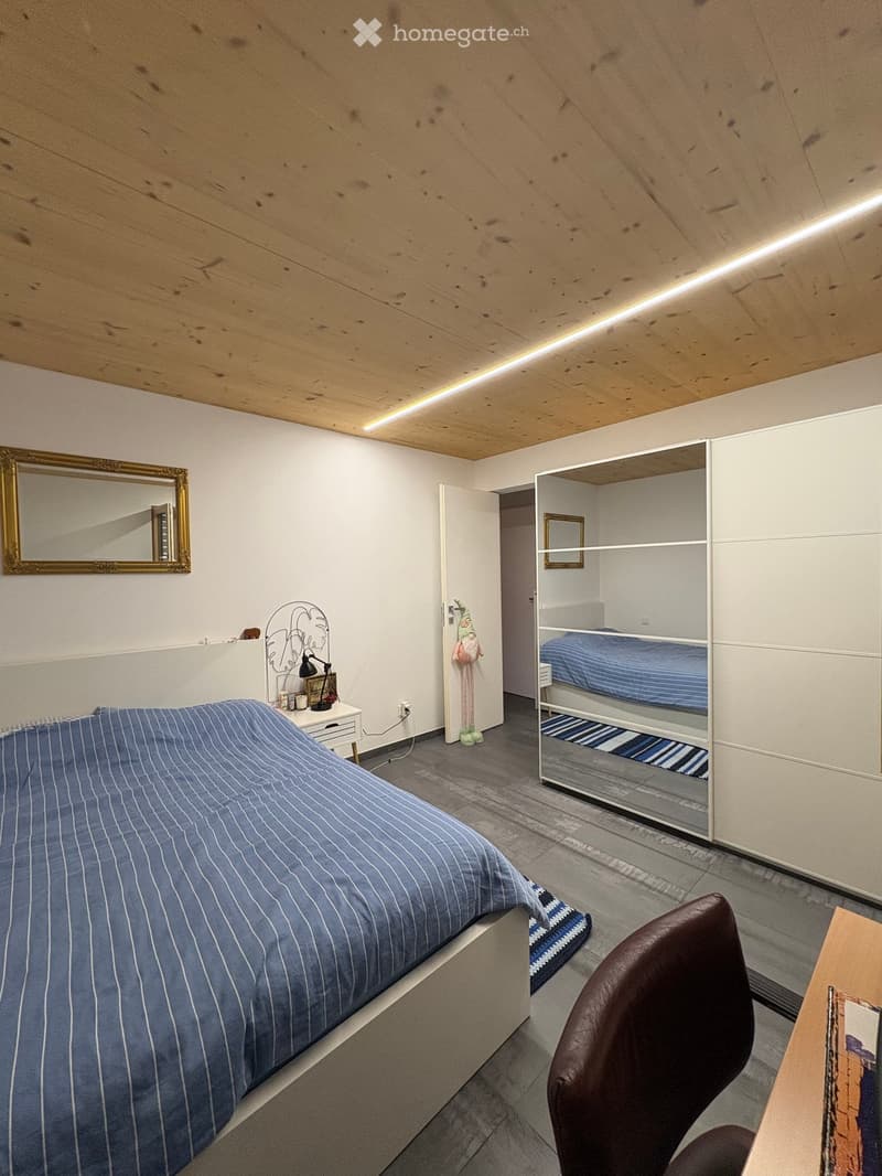 Cozy bedroom with a large bed, a chair, a small table, a mirror, a lamp, and a built-in wardrobe with a bunk bed inside.