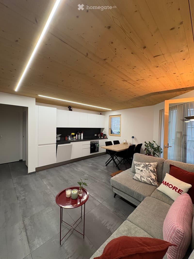 Modern living and dining area with a grey sofa, wooden dining table, kitchen area with white cabinets and black countertops, and a small round table with candles and a plant.