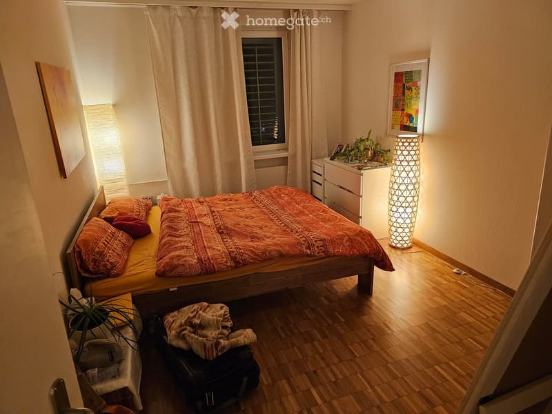 Cozy bedroom with wooden floor, bed with orange bedding, nightstand with lamp, small plant, and artwork on the wall.