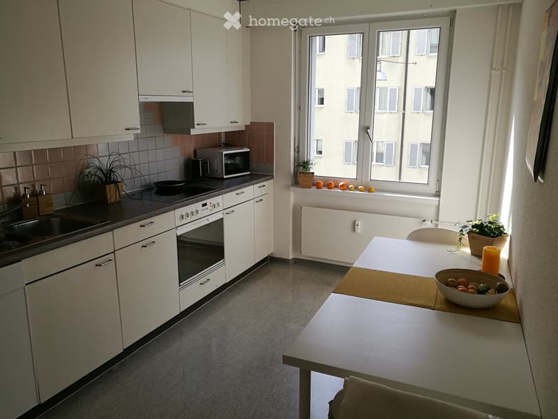 Modern kitchen with white cabinets, stainless steel appliances, a dining table with a bowl of fruit, and potted plants on the windowsill.
