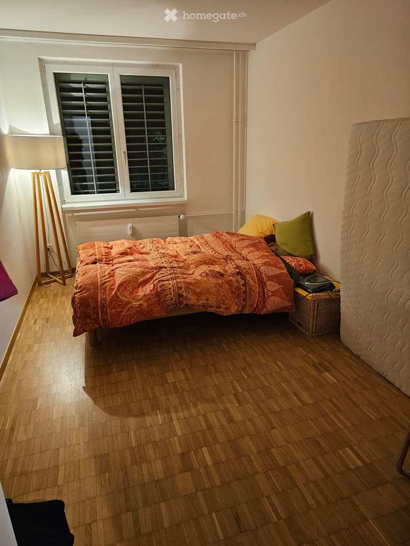 Cozy bedroom with wooden flooring, a bed with patterned bedspread and pillows, a nightstand with a lamp, and a window with blinds.