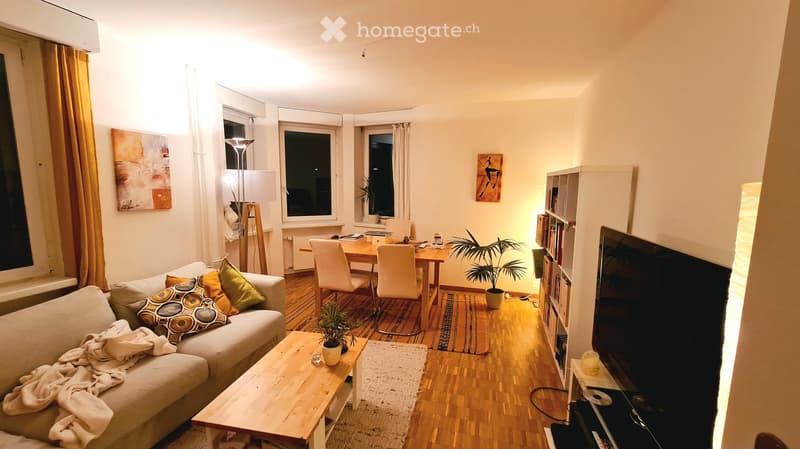 A cozy living room with a grey sofa, wooden coffee table, dining table with chairs, bookshelf, television, and several plants. The room features wooden flooring, large windows with curtains, and warm lighting.