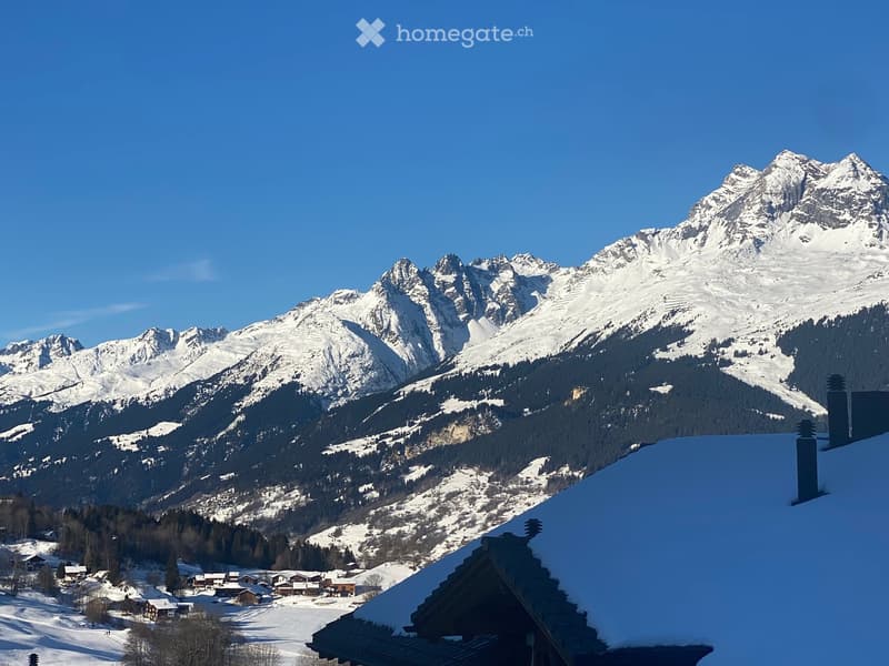 A panoramic view of snow-capped mountains and a village nestled in a valley, with a clear blue sky above.