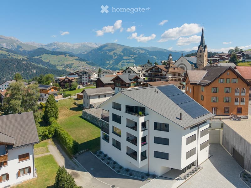Aerial view of a village with traditional houses, a church tower, and surrounding mountains.
