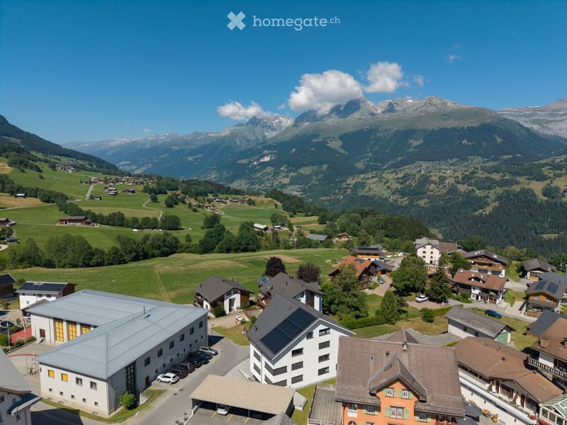 Aerial view of a village with houses, parking areas, and surrounding mountains.